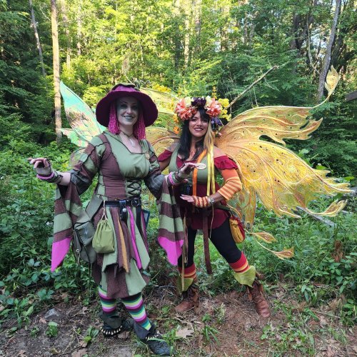 Two women in colorful, whimsical costumes stand in a lush green forest, surrounded by vibrant wings and floral accessories.