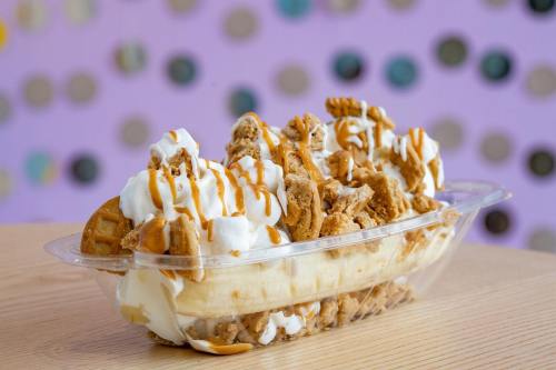 A dessert bowl with a banana, whipped cream, crumbled cookies, and drizzled caramel, set against a colorful background.