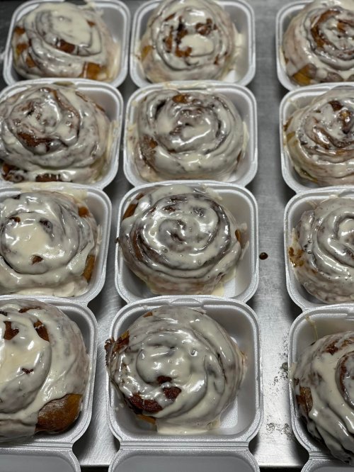A close-up of freshly baked cinnamon rolls with creamy icing, arranged in white takeout containers.