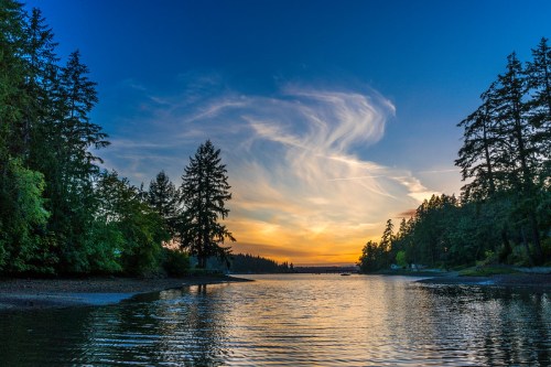 A serene river scene at sunset, with trees lining the banks and colorful clouds reflecting in the water.