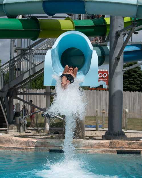 A person slides down a blue water slide, splashing into a pool below on a sunny day.