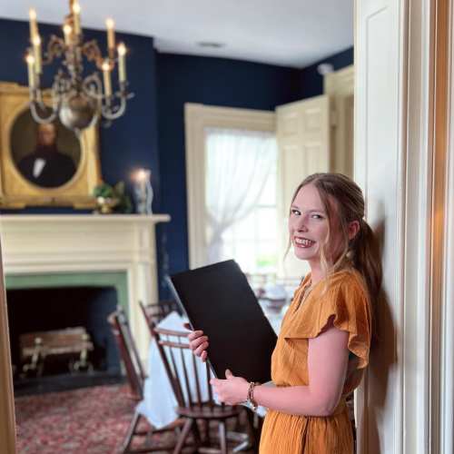 A smiling woman in a yellow dress stands by a doorway, holding a black folder in a beautifully decorated room.