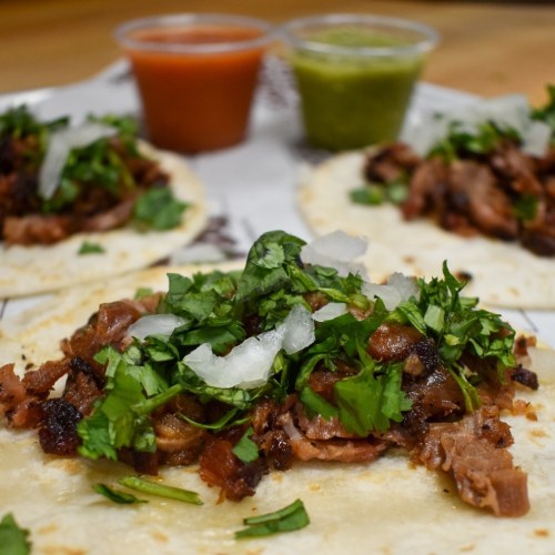 Three tacos filled with chopped meat, topped with cilantro and onions, served with red and green sauces on the side.