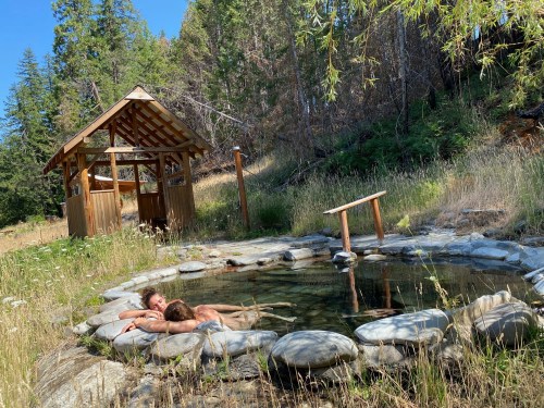 A serene outdoor hot spring surrounded by nature, with a wooden structure nearby and two people relaxing in the water.