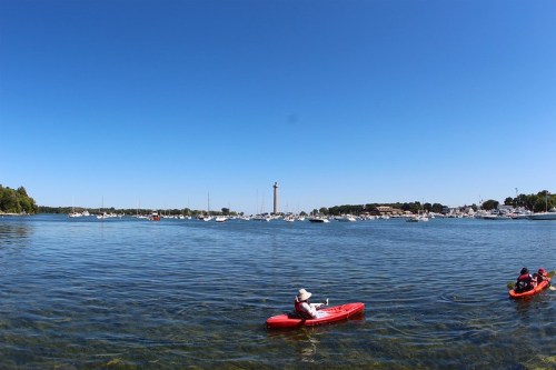 Two kayakers paddle in clear water near a marina with boats and a lighthouse under a bright blue sky.