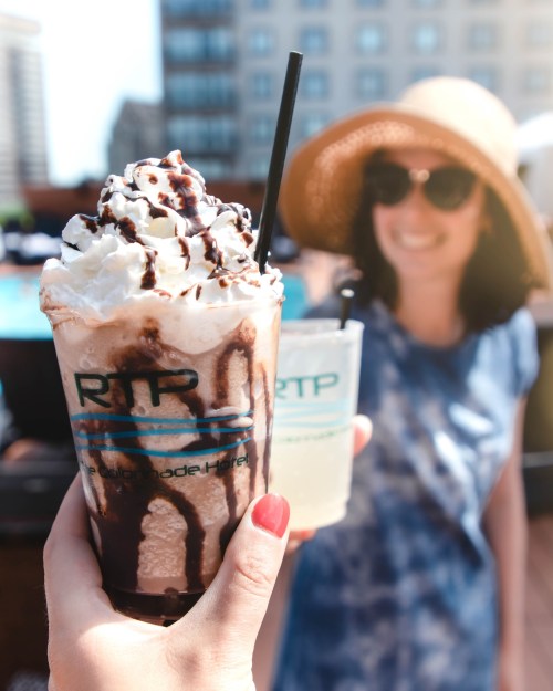 A hand holds a chocolate drink topped with whipped cream and chocolate drizzle, with a woman in a hat blurred in the background.