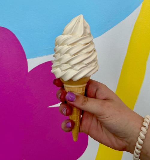 A hand holding a swirled soft serve ice cream cone against a colorful mural background.