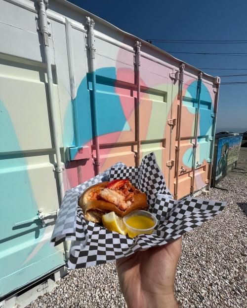 A hand holding a sandwich with fried fish and lemon, in front of a colorful mural on a shipping container.