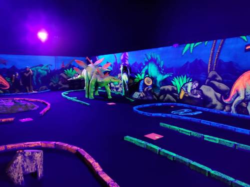 A vibrant mini-golf course with dinosaur decorations and glowing neon colors under purple lighting.