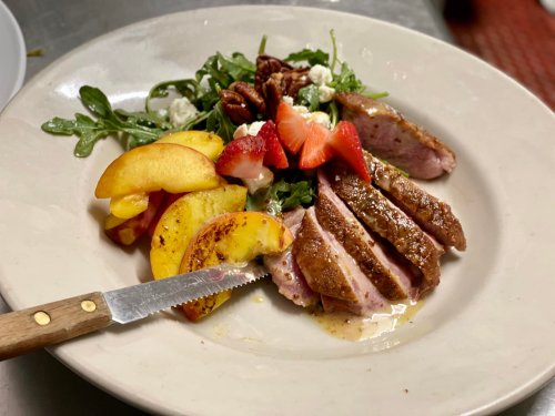 A plate of sliced duck breast with peaches, mixed greens, strawberries, pecans, and a knife on the side.