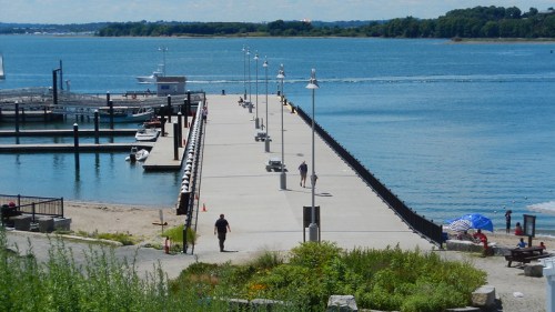 A sunny pier extends over calm water, with people walking and boats docked nearby, surrounded by greenery and beach.