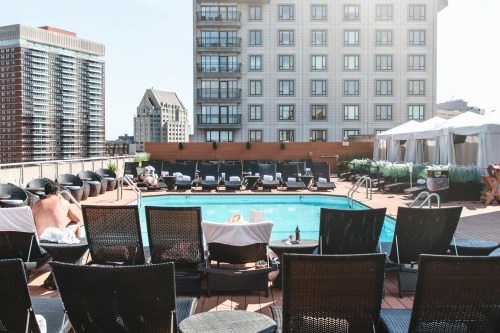 Rooftop pool area with lounge chairs, surrounded by city buildings and a sunny sky.
