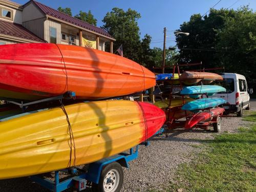 Colorful kayaks stacked on a trailer outside a building, with trees and a van in the background under a clear blue sky.