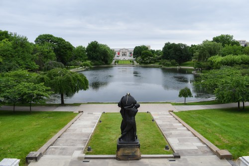 A serene park scene featuring a pond, lush greenery, and a statue in the foreground, with a building in the background.
