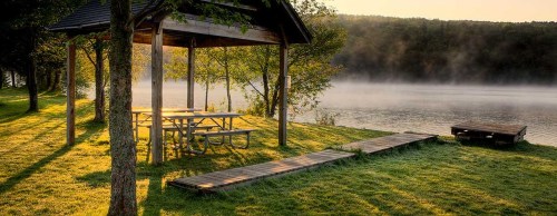 A serene lakeside scene with a wooden gazebo, picnic table, and mist rising from the water at sunrise.