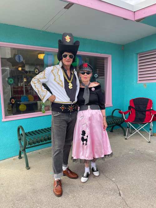 A man in a flashy outfit and sunglasses stands next to a woman in a poodle skirt and hat, both smiling outside a colorful building.
