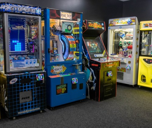 A row of colorful arcade machines, including a claw machine and games like Stacker and Road Trip, in a game room.
