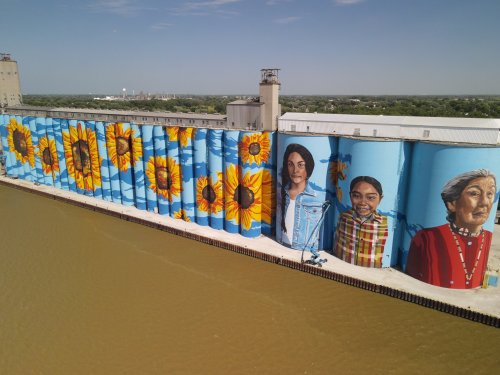Colorful mural on grain silos featuring sunflowers and portraits of three women against a blue background.