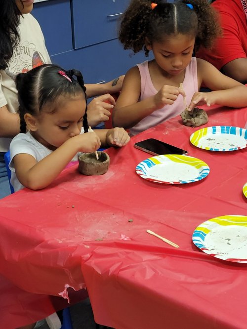 Two young girls focus on crafting with clay at a table, surrounded by colorful plates and a phone.