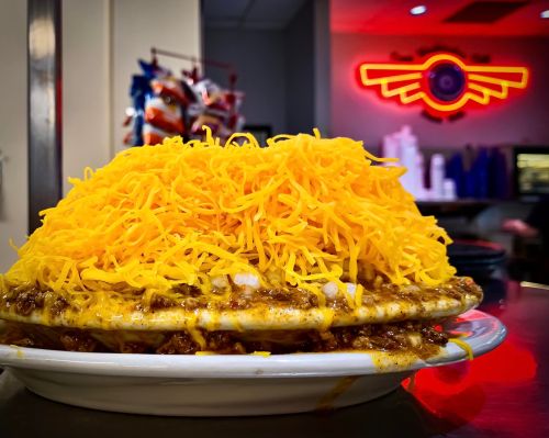 A large plate of chili cheese fries topped with a mound of shredded yellow cheese, set in a diner with neon lights.