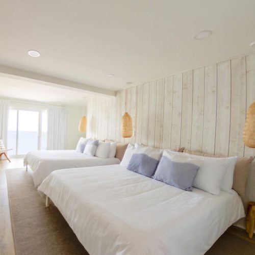A bright, airy bedroom with two beds, light wood paneling, and large windows overlooking a scenic view.
