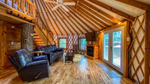 Cozy interior of a yurt with wooden beams, a fireplace, leather seating, and large windows letting in natural light.