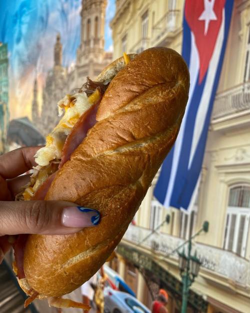 A hand holding a sandwich with layers of meat and cheese, set against a colorful backdrop featuring a Cuban flag.