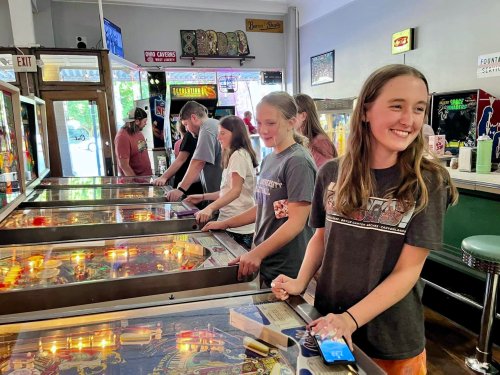 A group of people enjoying pinball machines in a retro arcade, smiling and engaged in the game.