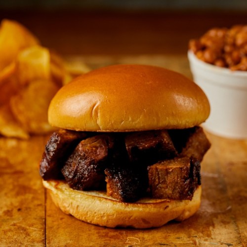 A close-up of a sandwich with chunks of meat on a soft bun, accompanied by chips and a small cup of beans.