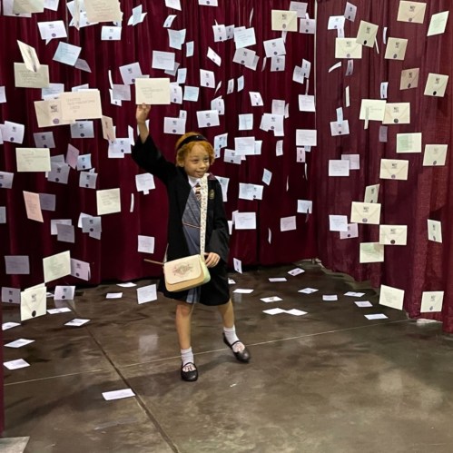 A child stands in a curtain-draped space, surrounded by floating notes and cards, holding one up with a smile.