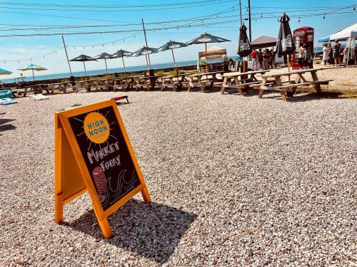 A sandy outdoor area with picnic tables, umbrellas, and a sign for "High Moon" market by the beach on a sunny day.