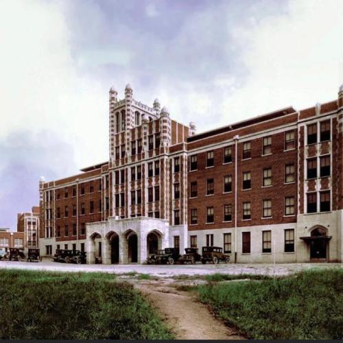 Historic brick building with ornate architecture, surrounded by grass and vintage cars parked in front.