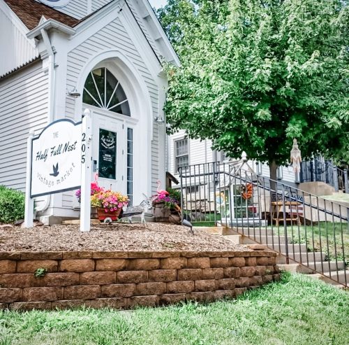A charming vintage shop with a sign, colorful flowers, and a tree in front of a quaint building.