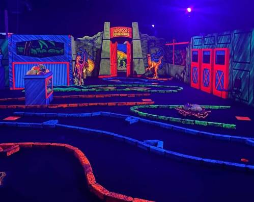 A vibrant indoor mini-golf course themed around dinosaurs, featuring glowing pathways and colorful decorations.