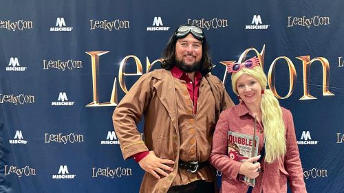 A man and woman in costumes pose together at LeakyCon, with a backdrop featuring the event's logo.