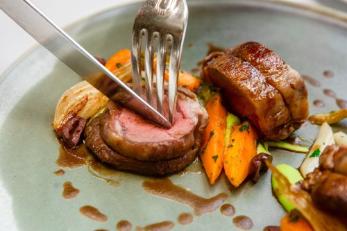 A close-up of a gourmet dish featuring sliced meat, carrots, and sauce, with a fork and knife cutting into the meat.