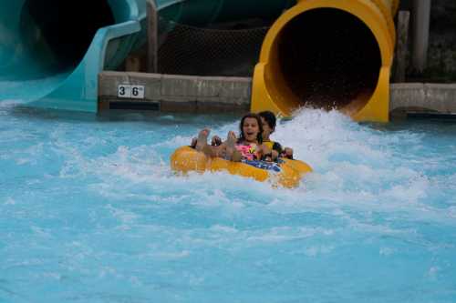 Two children joyfully ride a yellow inflatable tube down a water slide, splashing through the blue water.