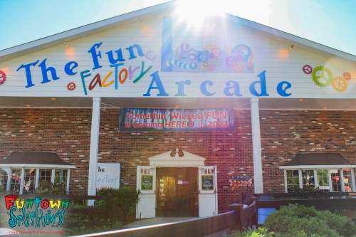 Brightly colored facade of "The Fun Factory Arcade" with a sunny sky and a sign announcing upcoming attractions.