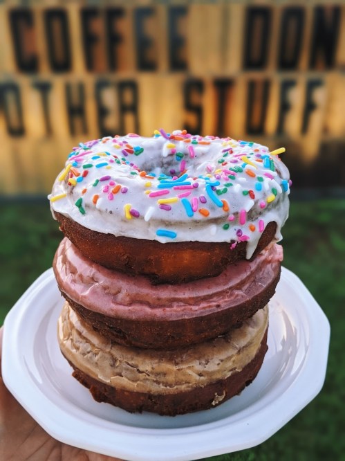A stack of three colorful donuts on a white plate, topped with frosting and sprinkles, against a rustic background.