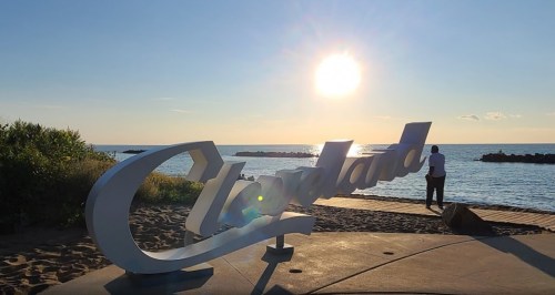 A person stands by a large "Cleveland" sign at the beach, with the sun setting over the water in the background.