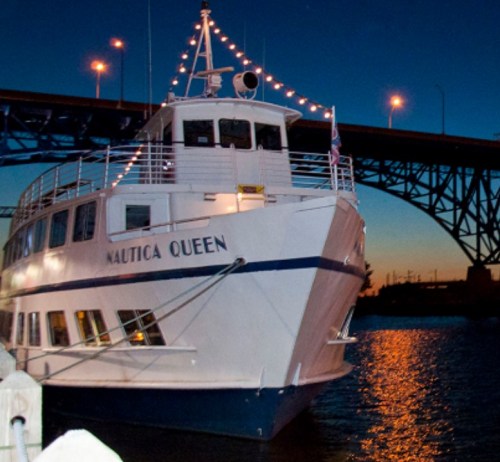 A lit-up boat named "Nautica Queen" docked under a bridge at dusk, reflecting on the water.