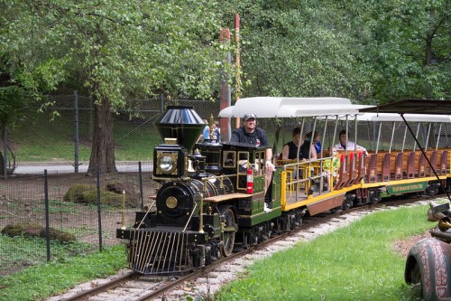 A miniature steam train with passengers travels along a track in a green park surrounded by trees.
