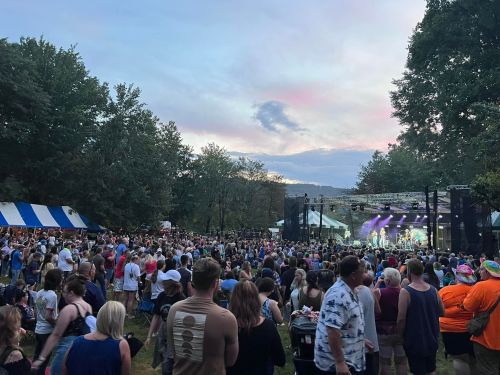 A large crowd enjoys a live music performance at an outdoor festival during sunset, with colorful tents in the background.
