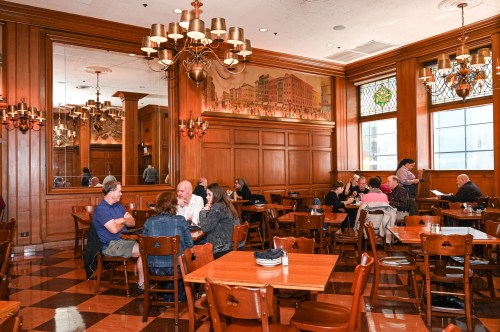 A cozy restaurant interior with wooden decor, tables occupied by diners, and large windows letting in natural light.