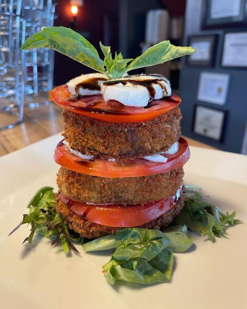 A stacked dish featuring crispy layers of fried food, fresh tomatoes, and a basil garnish on a bed of greens.
