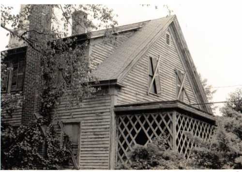 A weathered, two-story house with a steep roof, wooden siding, and overgrown vegetation around it.