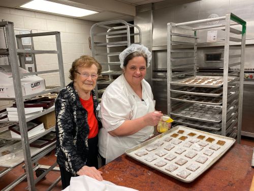 Two women in a kitchen, one wearing a chef's coat, preparing baked goods on a tray with racks of cookies in the background.
