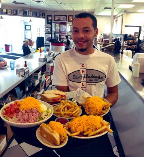 A smiling server holds a tray with a large salad, fries, and two loaded hot dogs at a diner.