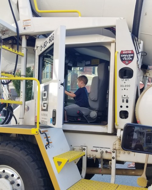 A young boy sits in the driver's seat of a large concrete mixer truck, looking focused and engaged.