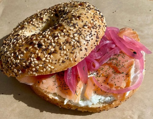 A sesame bagel filled with cream cheese, smoked salmon, pickled red onions, and dill on a brown paper background.